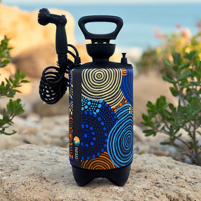 A black portable shower bottle featuring a vibrant abstract pattern of blue orange and yellow concentric circles and dots stands on light-colored rocks with a coiled hose and shower nozzle against a blurred blue ocean backdrop