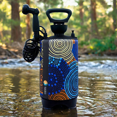 A portable shower with a vibrant patterned sleeve displaying concentric circles, scattered dots, and wavy lines in blue, yellow, and orange stands in a shallow river in a sun-dappled forest, featuring a coiled black hose and multi-setting nozzle