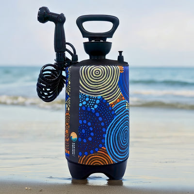 Product photo of a Portable Shower unit featuring a vibrant Aboriginal art-style pattern with blue, yellow, and orange concentric circles and dots, positioned on wet sand at the ocean's edge with gentle waves in the background
