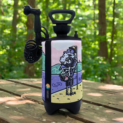 Photograph of a black portable pressure shower with a coiled hose and multi-setting showerhead nozzle, featuring a colorful sleeve with a melting ice cream skeleton holding a watermelon drink on a beach with purple water and a pink sky, standing on a wooden picnic table in a green forest setting