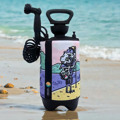 A black portable shower with a coiled hose and vibrant cartoon illustration of a skeleton with a foamy beard holding a cup stands on a wet sandy beach with a turquoise ocean and gentle waves in the background
