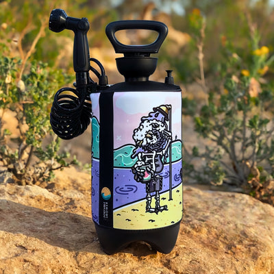 Photograph of a portable shower system with a black nozzle and coiled hose on a rocky outdoor surface, featuring a colorful cartoon wrap depicting a sudsy skeleton holding a watermelon drink on a vibrant beach with a purple ocean and pink sky