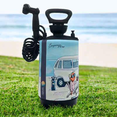A Benjamin Ross illustrated portable shower, light blue and black with a coiled hose and sprayer, stands on grass featuring a vibrant cartoon of a white VW camper van and a person in a folding chair on a sandy beach backed by a blue ocean