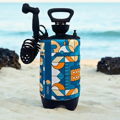 A product photograph of a portable shower unit featuring a vibrant blue, orange, and white geometric pattern, a black shower head and coiled hose, standing on a sandy beach with the ocean and dark rocks in the background