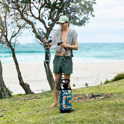 A man in a light green cap, grey towel, and olive green swim shorts prepares a portable shower with a vibrant blue, orange, and white geometric pattern on a grassy area with gnarled trees overlooking a sandy beach and turquoise ocean