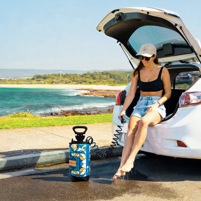 A woman in a black bikini and denim shorts rinses her bare feet with a colorful blue and orange portable shower, seated in a white car's open trunk, against a backdrop of a turquoise beach and rocky coastline