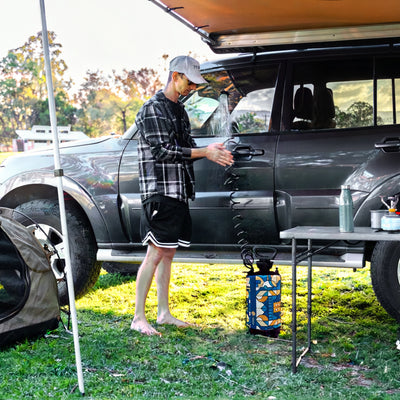 A man with a plaid shirt and black shorts showers his hands beside a grey SUV with an orange awning at a campsite, featuring a blue and orange patterned portable shower pump on the green grass