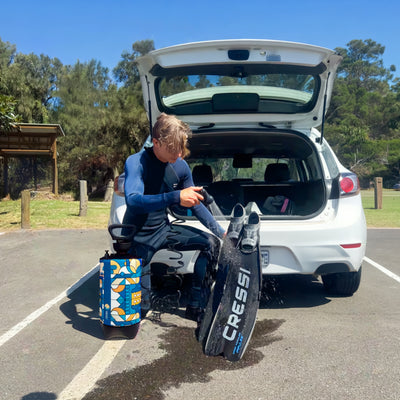 Photography of a young man in a blue wetsuit rinsing black Cressi diving fins with a colorful geometric patterned portable shower at a sunny beach parking lot next to a white car