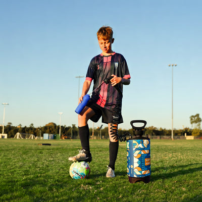 A young male soccer player in a black and pink striped jersey stands on a bright green field with one foot on a colorful "Deploy" soccer ball, holding a blue water bottle and a hose connected to a vibrant blue and orange patterned cleaning device, with stadium floodlights in the background