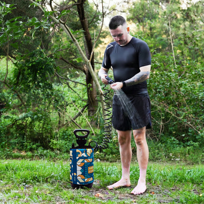 Photograph of a man in black athletic wear showering outdoors in a leafy forest environment using a portable pressure shower with a vibrant blue orange and beige geometric design and a coiled black hose