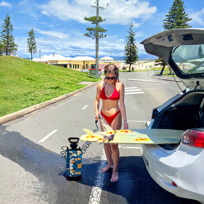 Photography of a woman in a red bikini washing a yellow surfboard with red star decals beside a white car and a blue geometric portable shower on a sunny coastal road with Norfolk Pines, yellow buildings, and distant sand dunes