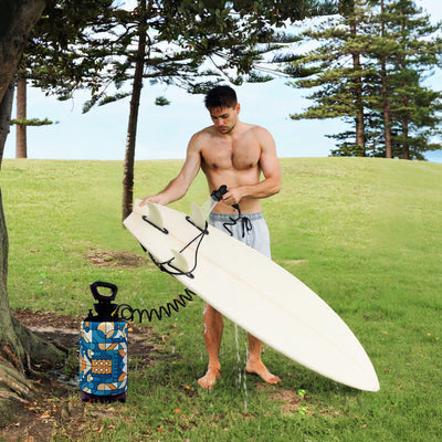 Photograph of a shirtless man cleaning a white surfboard with black fins using a portable blue and orange patterned shower unit under tall coastal pine trees on a grassy hill