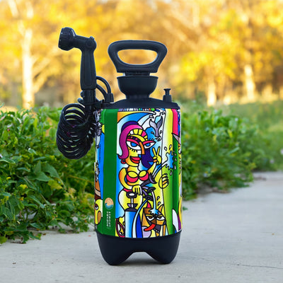 Photograph of a portable shower featuring a vibrant pop-art graphic of a stylized woman in a bikini making a peace sign alongside a dog and a whale tail, set on a concrete path with lush green bushes and blurred golden autumn trees