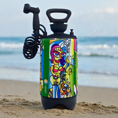 A portable shower with a vibrant Cubist-style artwork wrap depicting a woman in a pink bikini, a dog, and surfing elements stands on a sandy beach against a blurry blue ocean with its black coiled hose visible