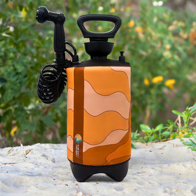 Photograph of a portable shower with an orange, peach, and brown wavy abstract pattern, featuring a black handle, coiled hose, and "Portable Shower" logo, sitting on sand against a lush green background with yellow flowers