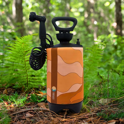 A portable camping shower with an orange wavy pattern, black coiled hose, and multi-setting nozzle stands on a forest floor amidst green ferns and trees