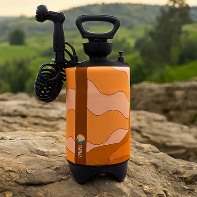 Product photo of a portable shower system featuring an orange, light orange, and brown wavy pattern sleeve, a black shower head, and a coiled hose, resting on textured rocks with blurred green hills in the background, displaying a "Portable Shower" brand label with a blue wave logo