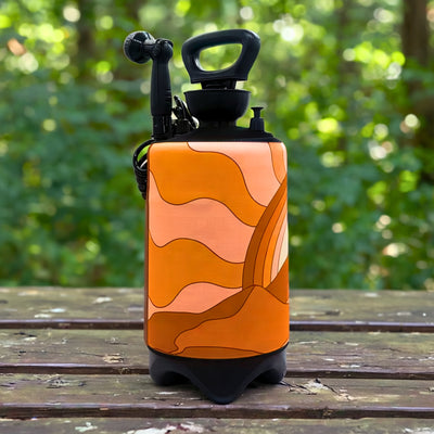 A portable pressurized camping shower featuring an orange, peach, and brown abstract wavy design with a black pump handle and nozzle sitting on a weathered wooden picnic table in front of a blurred green forest