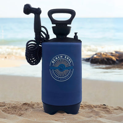 Product photo of a dark blue Beach Soul Portable Shower Pro with a prominent sun and wave logo, featuring a black handle and coiled hose, standing on a bright sandy beach against a blurred ocean backdrop