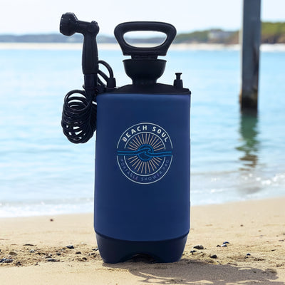 Product photo of a navy blue Beach Soul Portable Shower Pro with a black handle and coiled hose sitting on a sunlit sandy beach with the tranquil blue ocean and a pier post in the background