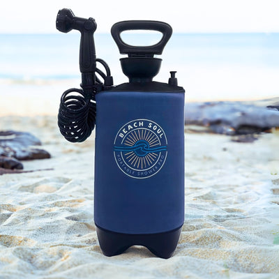 Product photograph of a dark blue Beach Soul Portable Shower Pro unit with a white circular sun and wave logo, black handle, and coiled hose standing on a bright sandy beach against a blurred ocean horizon