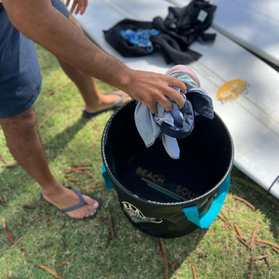 A person in blue swim shorts and flip-flops places light-colored swimwear into a black collapsible "BEACH SOUND" bucket with turquoise handles, beside a white surfboard on green grass