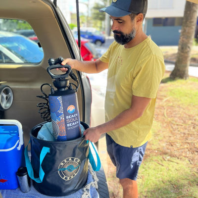 A bearded man in a yellow shirt and blue cap places a Beach Soul portable shower with vibrant blue and orange patterns into a black Beach Soul collapsible bucket in a car trunk next to a blue cooler outdoors