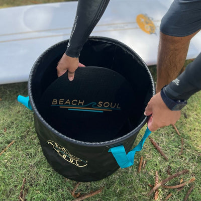 A person in a black wetsuit sleeve inserts a black circular mat with a "BEACH SOUL" logo into a black "BEACH SOUL" bucket featuring turquoise handles, set on green grass with scattered pine cones and a white surfboard with a yellow graphic in the background