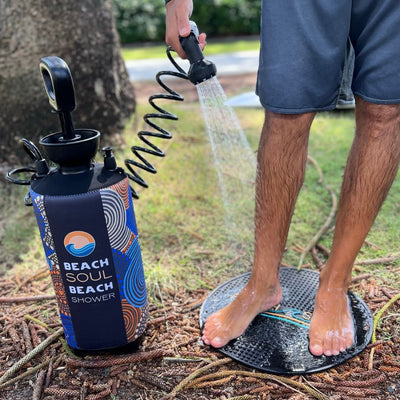 A person washes their wet feet on a black textured mat using a portable Beach Soul Beach Shower, featuring a vibrant blue and orange patterned sleeve, set on pine needles and grass