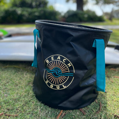 Black flexible bucket bag with teal handles showcasing a BEACH SOUL logo featuring a sun and wave design, placed on green grass with blurred white surfboards behind it