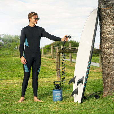 A young man in a black and teal wetsuit cleans a white surfboard with black fins and blue stripes using a portable blue Beach Soul pressure shower in a grassy outdoor area next to a wooden fence