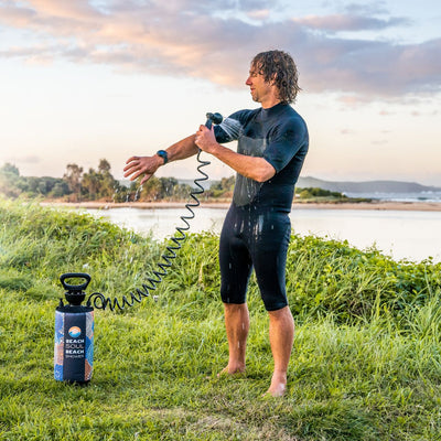 Photography of a man in a black wetsuit with wet curly hair rinsing his arm with a portable Beach Soul Beach Shower, standing on green grass by a calm body of water at sunset