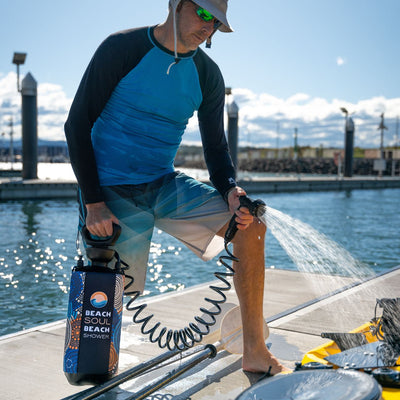 A man in a blue rash guard and patterned board shorts rinses his leg with a black and colorful Beach Soul portable shower on a sunny wooden dock by sparkling blue water with yellow kayaks nearby