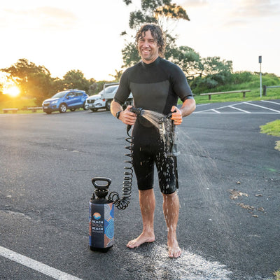Photograph of a smiling man in a black wetsuit rinsing off with a portable "Beach Soul Beach Shower" featuring colorful circular patterns in a sunlit parking lot with trees and cars in the background