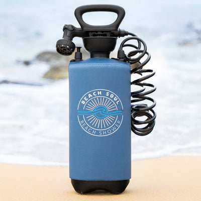 A dark blue Beach Soul portable shower with its brand logo, black coiled hose, and showerhead rests on a sandy beach near the ocean's gentle waves