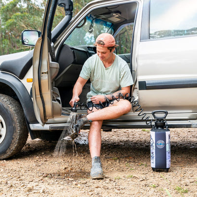 A man in a peach cap and "Assembly Label" t-shirt sits on the doorframe of a silver SUV on a dirt track, cleaning his muddy boots with a portable pressurized washer that features a distinctive black and white illustrated sleeve