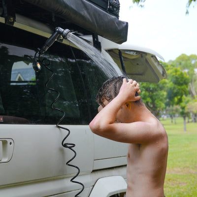 A shirtless man showering outdoors under a black portable coiled shower head attached to a white camper van with a black roof awning in a bright green park