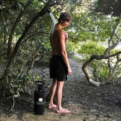 Photograph of a young man with wet hair and black shorts showering outdoors using a portable black Beach Soul shower unit with a colorful logo, water spraying, surrounded by lush green foliage and visible tree roots on a sandy path