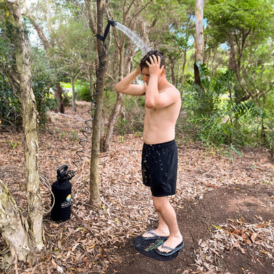 A man showering outdoors in a wooded area with a portable "BEACH SHOWER" system featuring a black canister and coiled hose, standing on a branded black mat as water sprays over him