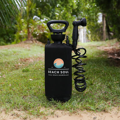 A black Beach Soul portable beach shower with a coiled hose and an orange and blue wave logo stands on green grass with a sandy edge in a lush outdoor setting