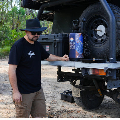 A man in a black hat and sunglasses stands by an overlanding ute in sunny bushland, alongside a vibrant blue and orange 'Beach Soul' portable shower and a small cast iron pan