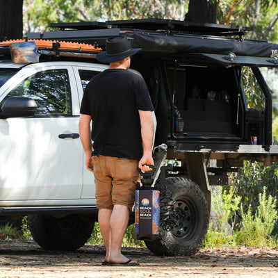 A man in a black hat stands beside his white off-road pickup truck with a black roof rack and shovel, holding a portable Beach Soul Beach Shower with a colorful wave and dot pattern in a natural wooded setting