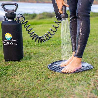 Photograph of a person in black wetsuit pants rinsing their bare feet on a black textured mat with a Beach Soul portable shower tank featuring a yellow sun and blue wave logo on a grassy beach shore