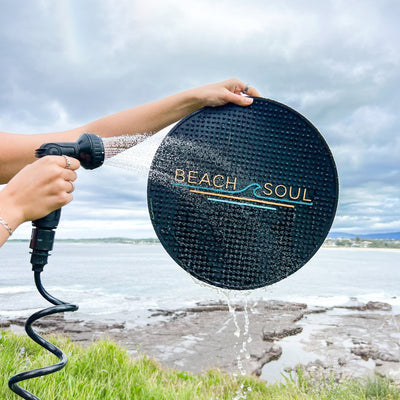 A person's hands holding a black hose nozzle spraying water onto a black textured circular "Beach Soul" mat featuring a teal and orange wave logo, set against a background of a rocky coast and ocean under a cloudy sky